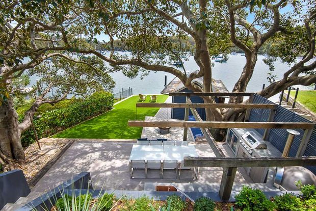 Elevated view of a landscaped outdoor entertainment area beneath mature trees, with a paved terrace, outdoor dining table, timber pergola structure, built-in outdoor kitchen or barbecue, manicured lawn, and glimpses of water beyond.