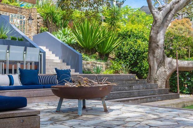 An outdoor landscaped courtyard or terrace with layered stone paving, dark stone retaining walls and steps, lush tropical plantings, a built-in blue-and-white cushioned seating area, and a shallow sculptural fire bowl. A stone-clad wall and decorative glazing are visible behind the seating.