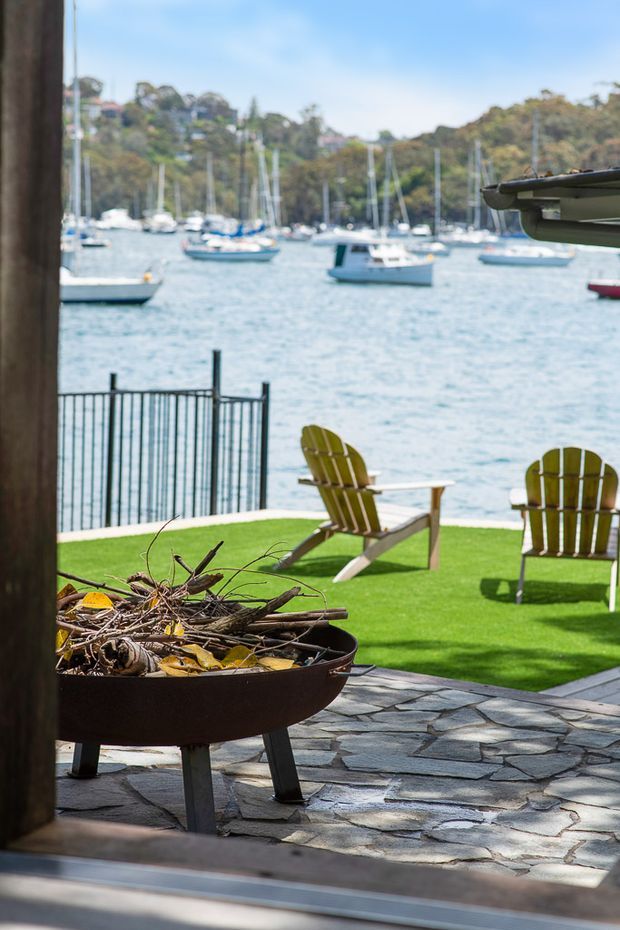 A waterfront outdoor entertaining area viewed from a building opening, with a dark stone-paved patio, circular fire pit, artificial or closely clipped green lawn, Adirondack-style chairs, and a metal railing overlooking a marina and wooded shoreline. A partial roof or awning is visible at the upper right.