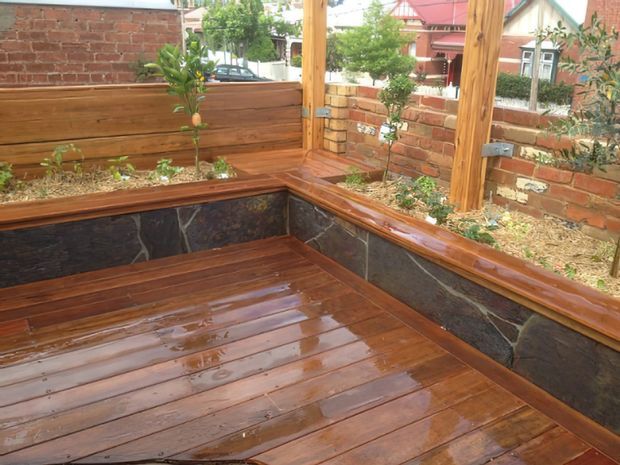 A compact outdoor courtyard or deck area with a freshly finished timber deck, built-in timber bench seating and raised planter beds. Dark natural-stone wall cladding runs below the benches, while timber posts and horizontal fencing frame the space. Brick boundary walls and neighbouring residential buildings are visible beyond.