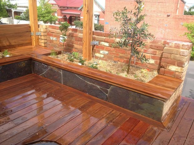 Outdoor timber deck with built-in corner bench seating and raised brick-and-stone planter walls. The space features warm polished timber decking and bench tops, dark natural stone wall insets, exposed timber posts, and brick garden walls with small planted trees. Residential buildings are visible beyond the courtyard-like outdoor area.