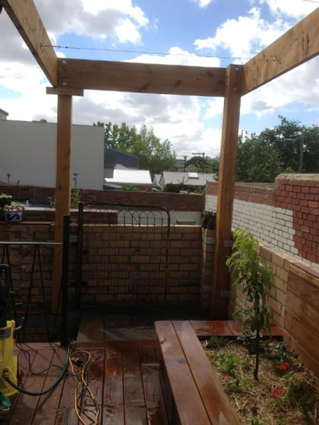 A small urban outdoor courtyard or terrace under construction, featuring a timber pergola frame, raised brick planter walls, timber decking, built-in bench seating, and a narrow garden bed. The space overlooks neighbouring rooftops and is brightly lit by daylight.
