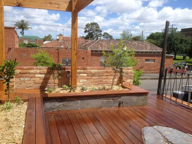 A timber deck or terrace with rich reddish-brown wood decking, raised built-in planter seating, and a timber pergola structure. A low stone-faced retaining wall and planted garden border the space, with a suburban brick building and tiled roof visible beyond the railing.