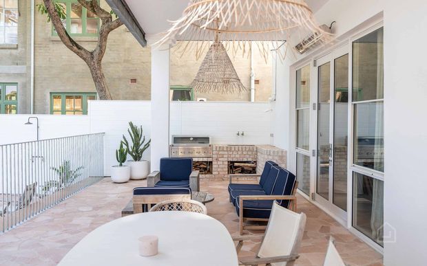 A bright covered outdoor terrace adjoining a home, furnished with a round dining table, lounge chairs and a built-in brick barbecue/fireplace. The space has pale stone or terracotta-look paving, white walls and railings, extensive glass doors, and woven pendant lights, with a mature tree and potted plants adding an organic touch.