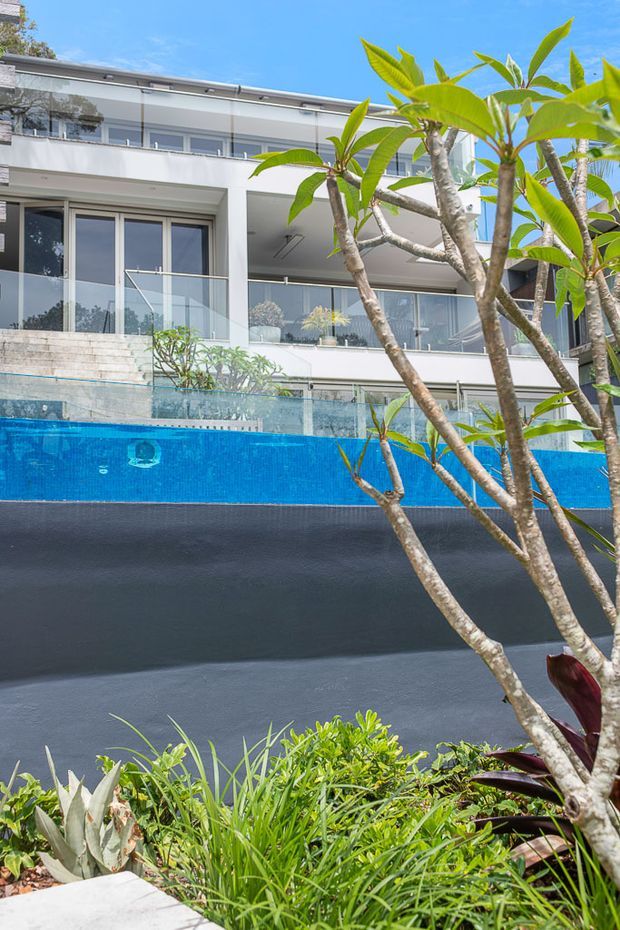 Exterior view of a contemporary multi-storey white house with expansive glazing, balconies and terraces overlooking a bright blue swimming pool. Tropical planting, including frangipani trees, occupies the foreground, with dark pool surrounds and transparent glass pool fencing.