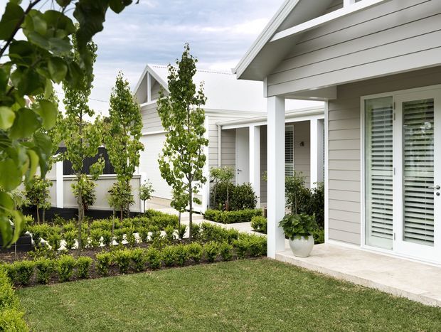 A bright exterior view of a contemporary residential development with multiple light-painted, horizontal-clad buildings, gabled roof forms, covered porches, and white-framed glazed doors with plantation shutters. A landscaped courtyard garden with clipped hedges, young trees, shrubs, and lawn connects the buildings.