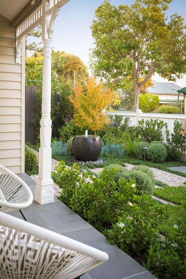 A bright, landscaped front veranda or porch beside a weatherboard house, featuring ornate white timber posts and fretwork, dark grey paving, woven outdoor lounge chairs, manicured hedges, layered planting, and a central bubbling water feature. A white picket-style fence and neighbouring roofline are visible beyond the garden.