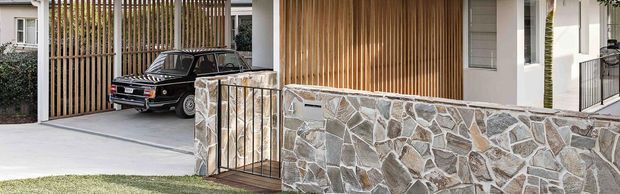 Contemporary residential front entrance and driveway featuring a pale rendered facade, vertical timber screening, a grey concrete driveway, a low irregular natural-stone boundary wall, and a black metal pedestrian gate. A parked vehicle is visible behind the screen.