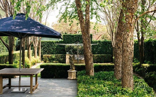 A landscaped outdoor courtyard or garden terrace with mature trees, dense clipped hedges, groundcover planting, and a paved stone patio. A timber outdoor dining table sits beneath a large dark umbrella, with a classical pedestal feature near the centre and a clothes-drying rack in the background.