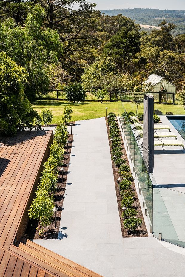 A landscaped outdoor pool area viewed from an elevated position, with a large pale paved walkway and terrace surrounding a glass-fenced swimming pool. Timber decking occupies the left and foreground, bordered by neatly planted hedges and garden beds. Poolside loungers and a closed outdoor umbrella sit beside the water, while a manicured lawn, trees, and a small pitched-roof outbuilding extend into the background.