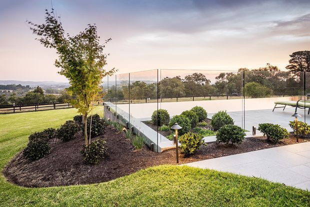 A landscaped outdoor residential setting featuring a fenced tennis court, manicured lawn, mulched garden beds with shrubs and young trees, and a paved terrace with outdoor seating. The scene overlooks a broad green valley beneath a warm evening sky.