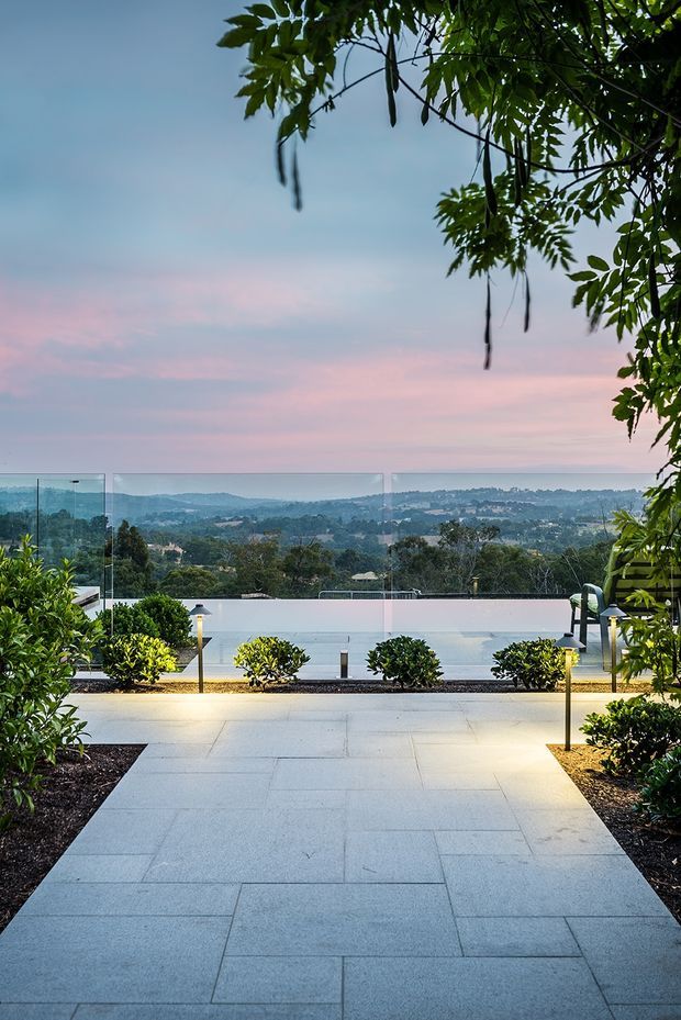 A landscaped outdoor terrace or poolside area with a broad pale stone-paved walkway leading toward a glass balustrade and panoramic rolling-hills views. Low clipped shrubs, garden beds, slender pathway lights, a bench, and overhanging leafy vegetation frame the scene beneath a soft pink-and-blue evening sky.