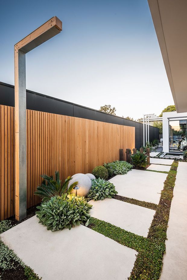 A modern landscaped side passage or courtyard walkway with large pale concrete stepping slabs separated by narrow strips of low groundcover. A tall vertical timber slat fence sits beneath a dark metal-clad wall, complemented by a sculptural timber-and-metal light fixture, drought-tolerant planting, spherical garden sculpture, and a glimpse of a contemporary house at the far end.