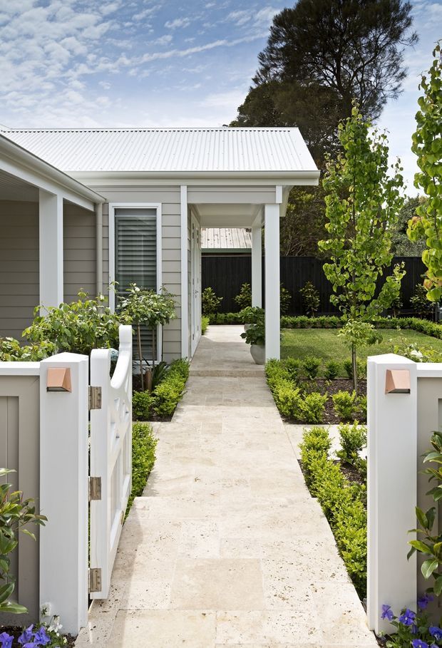 A landscaped front entry to a light-coloured single-storey home, featuring a white corrugated metal pitched roof, pale horizontal weatherboard cladding, white posts and trims, and a covered walkway leading to the entrance. A white pedestrian gate opens onto a pale stone-paved path, bordered by clipped hedges, lawn, young trees, and garden planting.