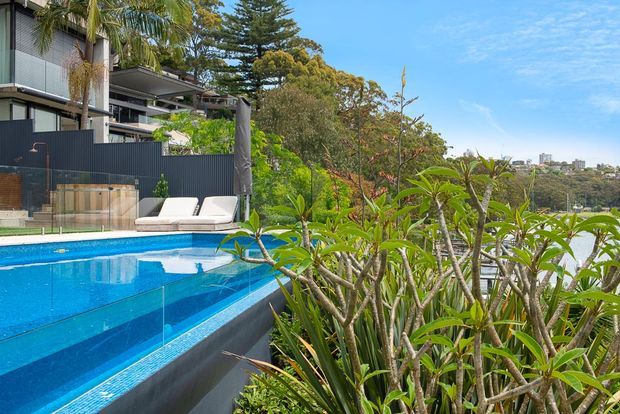 Bright exterior view of a contemporary multi-level residence with a vivid blue swimming pool in the foreground, landscaped tropical planting, and glass balustrades. The property overlooks a leafy urban landscape and distant buildings.