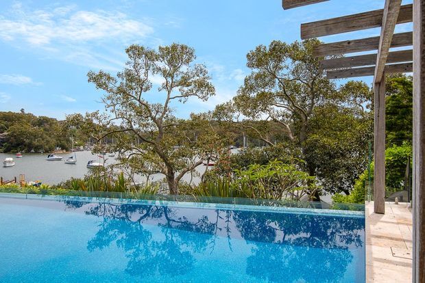 A bright outdoor view of a turquoise swimming pool overlooking a sheltered leafy waterfront inlet with moored boats. A timber pergola and pale stone-paved edge frame the right side, while native-looking vegetation and trees line the water beyond.
