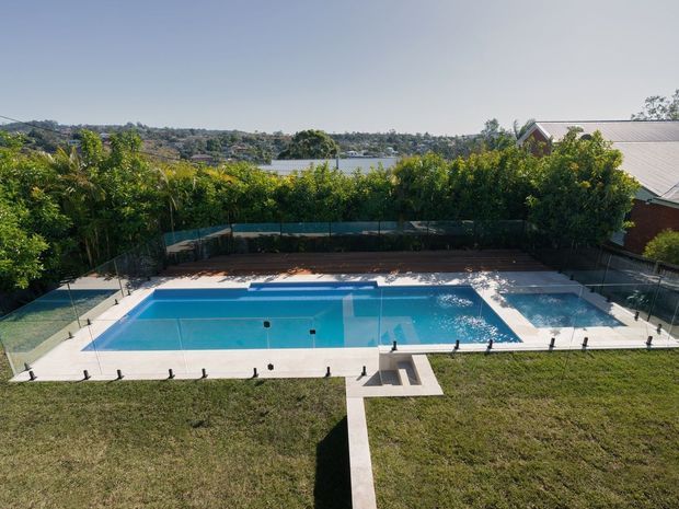 Aerial view of a landscaped residential pool area with a large rectangular blue swimming pool, pale paved surround, safety fencing, lawn, planted screening, and a straight concrete path leading to the pool. A lake or river and neighbouring homes are visible in the distance.