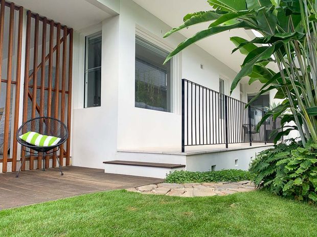 A contemporary residential exterior courtyard or terrace with white rendered walls, dark-framed windows, black vertical metal balustrades, and a timber privacy screen. A timber-look deck and stepping-stone path connect to a landscaped lawn with lush tropical planting. A small outdoor chair with a striped cushion sits beside the screen.