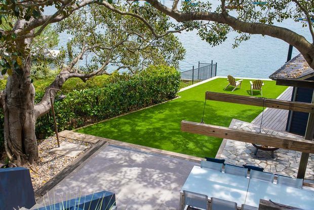 A landscaped waterfront outdoor area viewed from an elevated terrace, with a large mature tree, paved entertaining space, bright green lawn, outdoor chairs, timber-and-rope balustrade, stone paving, and a glimpse of a dark-clad structure at right. Water and a fenced edge are visible beyond the lawn.