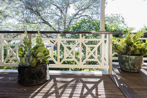 Sunlit timber deck or balcony with a decorative white geometric balustrade, dark timber handrail, and potted plants. Mature trees and greenery are visible beyond the railing, creating a leafy outdoor setting.