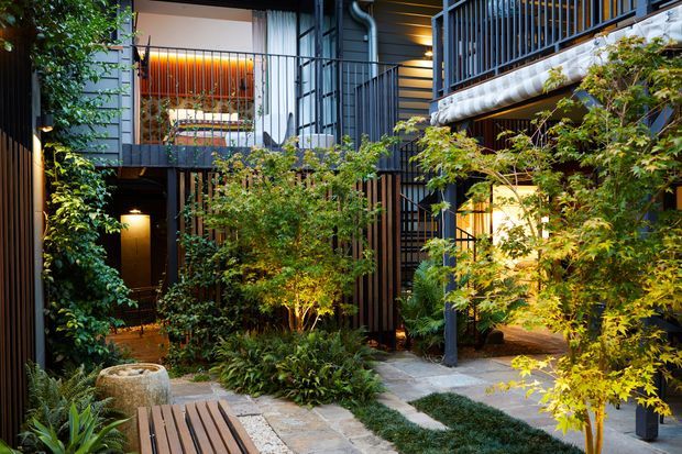 A lush landscaped courtyard enclosed by a multi-level contemporary building, with dark metal balcony railings, vertical timber screening, warm interior lighting, stone paving, planted garden beds, and a timber bench in the foreground. The courtyard has a calm, intimate, biophilic character.