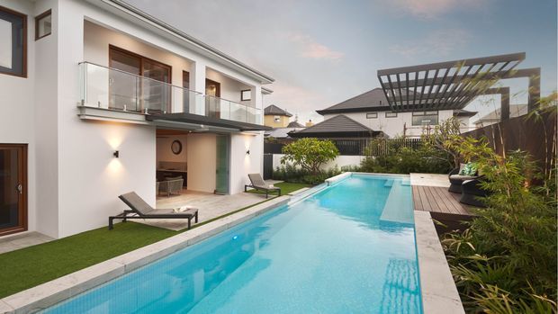 A contemporary multi-level residential home viewed from the pool courtyard, with a long turquoise swimming pool, tiled paving, glass-balustraded upper balcony, white rendered walls, dark-framed doors and windows, and a timber pool deck beneath a large slatted pergola. Landscaped planting borders the pool and outdoor lounge areas.