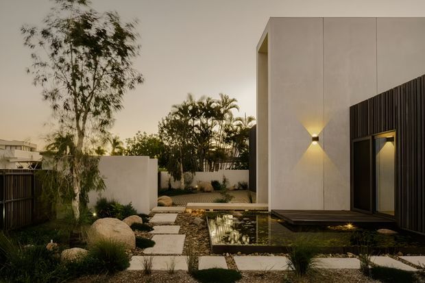 A landscaped contemporary residential courtyard at dusk, featuring a minimalist white rendered building with a tall dark vertical-slat entrance screen, warm wall lighting, a shallow reflective pond, stepping-stone paving, ornamental grasses, sculptural boulders and tropical planting.