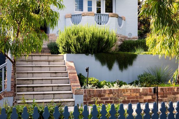 Bright exterior view of a raised residential entry, featuring broad tiled steps, layered landscaped garden beds, retaining walls, brick edging, and a white rendered or painted brick facade with a small balcony and decorative balustrade. Mature foliage frames the approach.