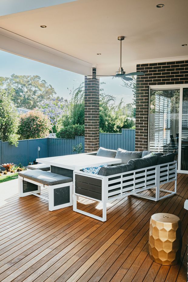A covered outdoor patio or terrace with a timber deck, white-framed built-in brick columns, and a large outdoor sectional dining lounge. The space includes a white rectangular table, grey cushions, a hanging pendant light, glass doors with blinds, and a blue privacy fence opening onto a landscaped garden.