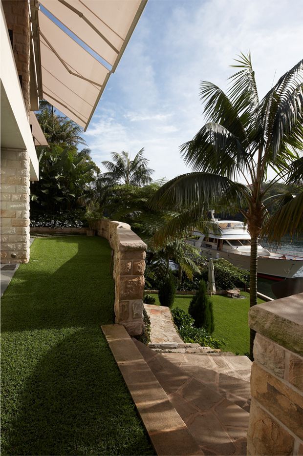 A tropical waterfront exterior viewed from a stone-clad terrace and landscaped garden. The scene includes broad stone-paved steps and retaining walls, manicured lawn, lush palm vegetation, a large boat below, and a retractable fabric awning attached to the building.