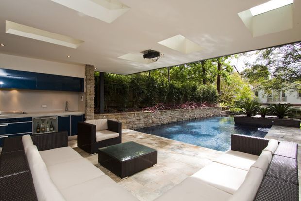 A covered outdoor entertaining area beside a swimming pool, featuring a built-in outdoor kitchen with dark blue cabinetry, extensive lounge seating, stone paving, a stone retaining wall, and multiple skylights in the roof. Dense greenery surrounds the pool and patio.
