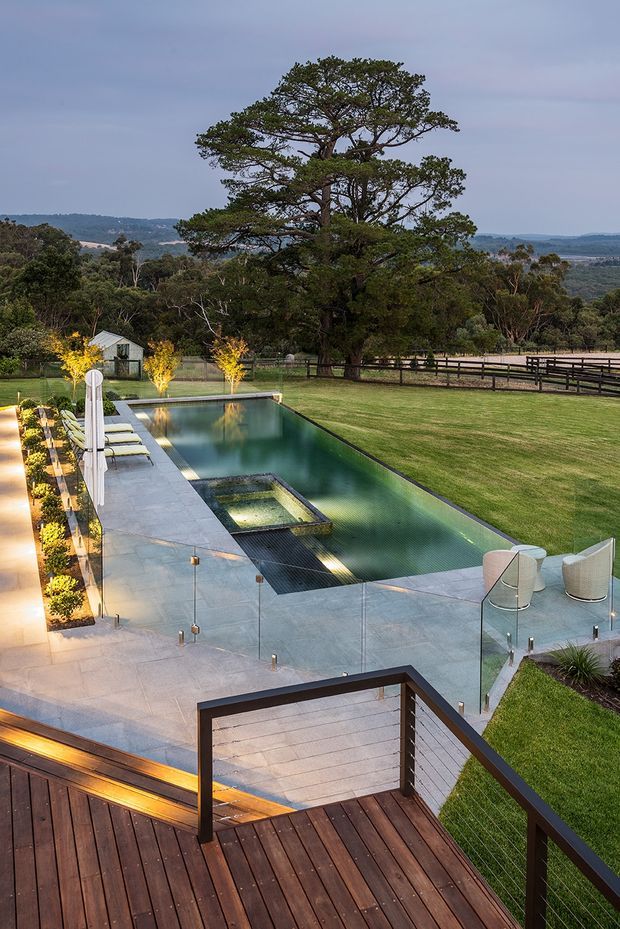 A landscaped rural property viewed from an elevated timber deck, featuring a long rectangular swimming pool with an integrated spa, pale stone paving, frameless glass fencing, and outdoor seating. Manicured lawn, mature trees, fencing, and distant rolling hills create a spacious countryside setting.
