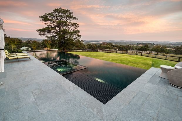 A contemporary outdoor pool terrace overlooking a broad rural or suburban landscape at sunset. The rectangular dark-tiled infinity-edge pool is surrounded by pale stone paving, with a spa or water feature at one end, outdoor seating, umbrellas, lawn, and a timber or dark metal boundary fence. A large mature tree and distant rolling landscape form the backdrop.