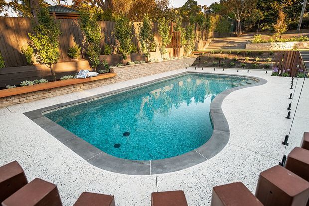 A landscaped residential backyard with a curved in-ground swimming pool surrounded by a pale speckled concrete terrace. The pool has a dark grey coping border, turquoise water, and integrated steps, with timber fencing, raised masonry garden beds, built-in timber bench seating, dense planting, and outdoor seating around the pool.