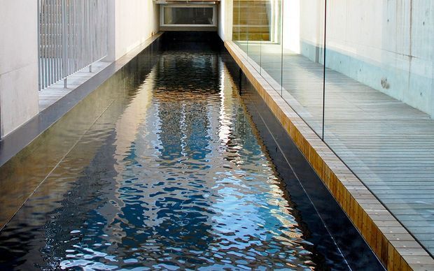 A narrow, elongated indoor lap pool or water feature runs through a modern architectural space. The pool is bordered by dark tiled edges and light tiled walkways, with vertical metal screening on one side and large glazed openings and pale concrete walls on the other. The composition is linear and minimalist, emphasizing reflective water and strong geometric lines.