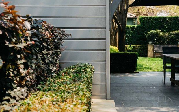 A landscaped outdoor patio beside a contemporary grey horizontally clad wall. Dark grey tiled paving leads through an open passage to a neatly manicured garden with clipped hedges, mature trees, and a timber outdoor dining table and chairs. The scene has a refined, structured garden design with strong natural greenery and neutral architectural finishes.