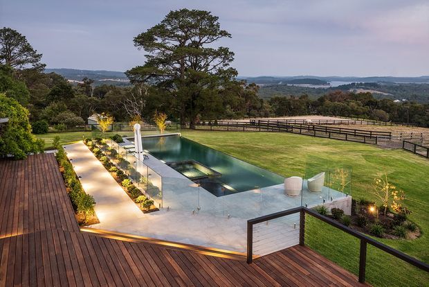 A landscaped outdoor entertaining area viewed from an elevated timber deck, featuring a long rectangular swimming pool with an integrated shallow spa or water feature, glass balustrades, illuminated garden paths, manicured lawns, and expansive rural or coastal views beyond. The setting is spacious and refined, with dark timber decking and contemporary poolside detailing.