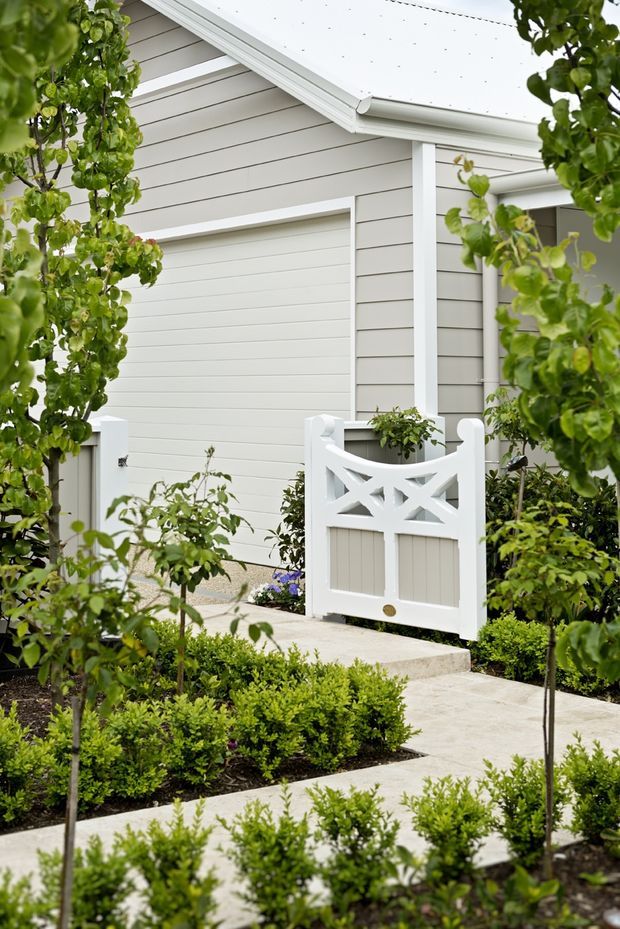 A landscaped residential side entrance beside a garage, featuring light grey horizontal weatherboard cladding, white trim, a white pitched roof edge, and a decorative white garden gate with lattice detailing. Pale concrete paths are bordered by neatly clipped low hedges, espaliered or young fruit trees, and ornamental planting.