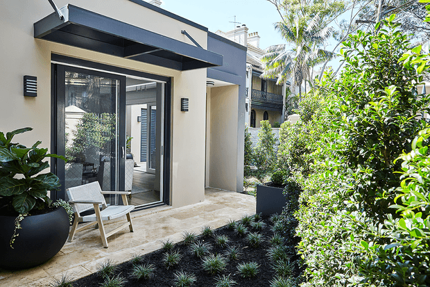 A contemporary residential courtyard or garden viewed from outside, featuring a pale rendered facade, dark projecting awning, large glazed sliding door, and a paved terrace with outdoor seating. Dense manicured planting, black planters, and a dark mulched garden bed frame the entry, with neighbouring residential architecture visible beyond.