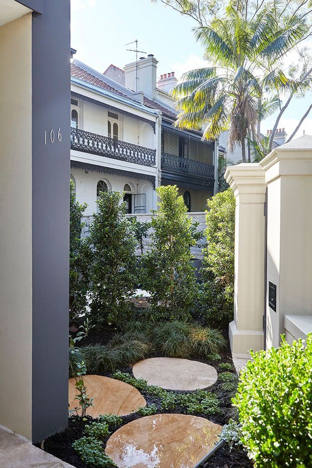 A landscaped residential courtyard or entry garden viewed between cream rendered walls, with large round stepping stones set through dark planting beds and clipped shrubs. The background shows ornate historic terrace-style homes with arched windows, decorative wrought-iron balconies, tiled roofs, and chimneys, framed by mature palms and greenery. A dark contemporary wall marked with the number 106 contrasts with the heritage architecture.