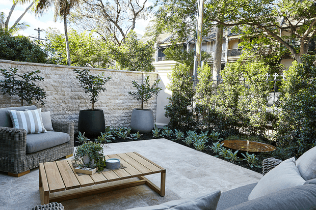 A refined outdoor courtyard terrace furnished with a low timber coffee table, woven lounge chairs and a pale upholstered sofa. Large concrete planters, layered greenery, a textured stone boundary wall and a water feature create a private, leafy entertaining space. The terrace is brightly lit with a calm, contemporary residential feel.
