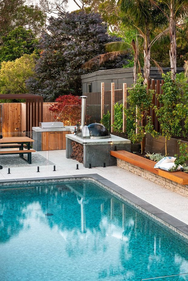 A landscaped residential pool area with a turquoise swimming pool, light aggregate paving, timber privacy fencing, raised planter beds, integrated timber bench seating, and an outdoor kitchen or barbecue zone. A compact pavilion-like structure and abundant mature planting surround the entertaining space.