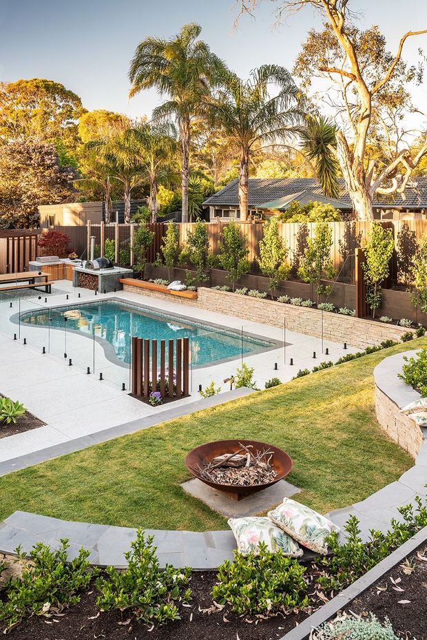 A landscaped residential backyard centred on a swimming pool, with pale concrete paving, frameless glass pool fencing, raised garden beds, clipped shrubs, lawn, a circular fire bowl, and a curved stone retaining wall. Tall palms and established trees surround a timber-fenced property with a tiled-roof house in the background.