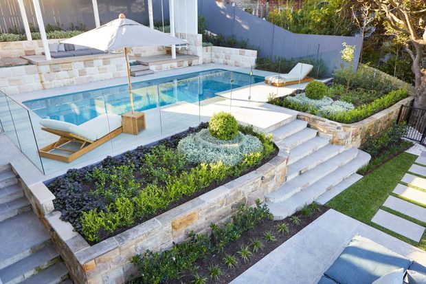 A landscaped residential pool area viewed from above, featuring a clear glass pool fence, rectangular swimming pool, pale stone paving and retaining walls, broad stone steps, raised garden beds, and sun loungers beneath a large umbrella. The design has a bright, refined contemporary outdoor setting with layered planting and strong geometric lines.