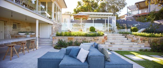 A bright, landscaped residential courtyard and outdoor entertaining area viewed from a lower terrace. The scene features a large blue outdoor sectional sofa in the foreground, a built-in outdoor bar or counter with stools at left, broad stone steps, planted garden beds, clipped shrubs, and a covered pavilion with an outdoor dining setting. Multi-level homes and balconies frame the space.