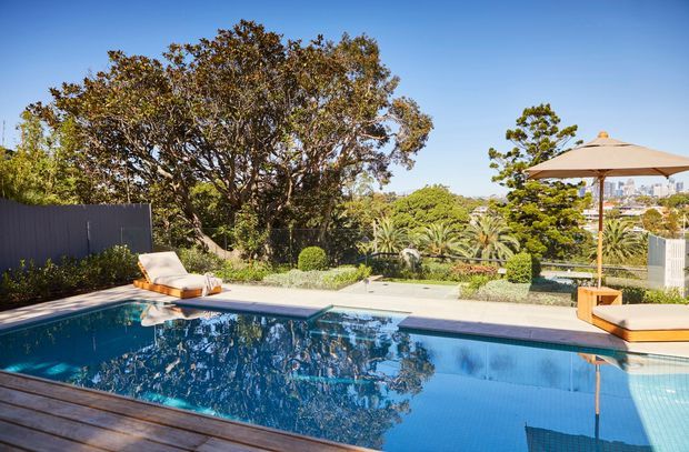Sunny landscaped residential pool area with a large rectangular swimming pool, timber decking in the foreground, lounge chairs, a cantilevered beige parasol, clipped garden planting, and a distant urban skyline visible beyond the property.