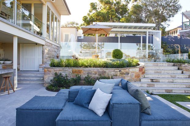 A bright landscaped outdoor entertaining area beside a contemporary multi-level home. The foreground features a large blue outdoor sectional lounge, while a raised stone planter with clipped greenery separates it from a dining area with a timber table and umbrella beneath a light pergola. Broad stone steps, frameless glass balustrades, extensive glazing, and layered garden terraces define the space.