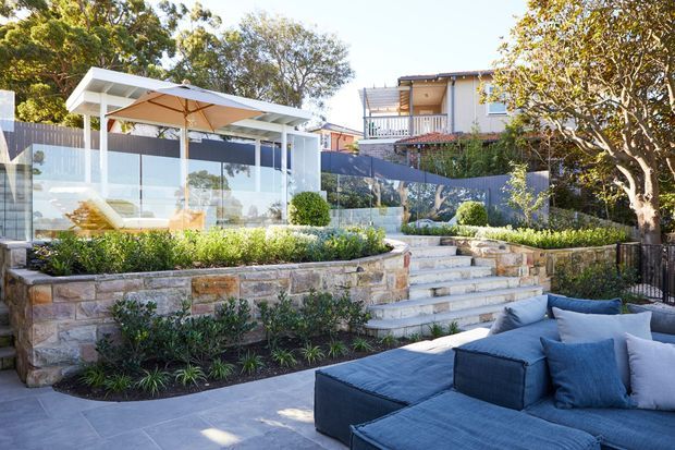A bright landscaped outdoor entertaining area with a large blue-grey sectional lounge on a paved terrace in the foreground. Stone retaining walls, stepped garden beds, clipped shrubs, and broad tiled steps lead up to a glazed pool or pavilion enclosure with a flat-roofed white structure and outdoor umbrella. A neighbouring residential building and mature trees are visible beyond the property.
