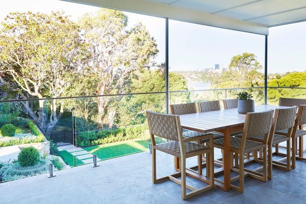 Bright covered outdoor dining terrace with a large timber dining table and woven chairs, enclosed by frameless glass balustrades and expansive glazing. The terrace overlooks landscaped gardens and a distant urban or waterfront view, with a clean, contemporary architectural setting.