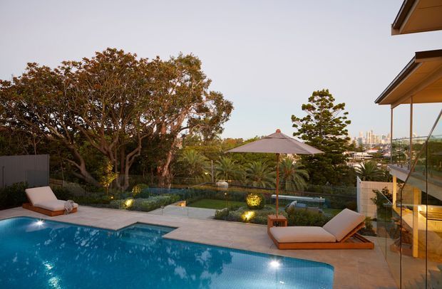 A landscaped outdoor pool area viewed from a contemporary home, featuring a blue swimming pool, paved pool deck, sun loungers, a large patio umbrella, planted garden beds, mature trees, and glazed balconies or living spaces overlooking the pool. A distant urban skyline is visible beyond the garden.